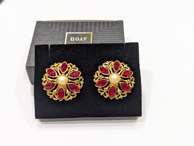 May include: A pair of gold tone earrings with a red flower design and a pearl center. The earrings are in a black velvet box with the word "AVON" printed on the box.