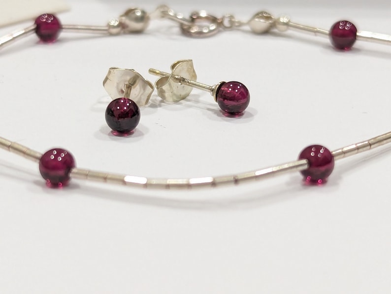 May include: A silver bracelet with five dark red gemstone beads and matching stud earrings. The bracelet is made of small silver beads.