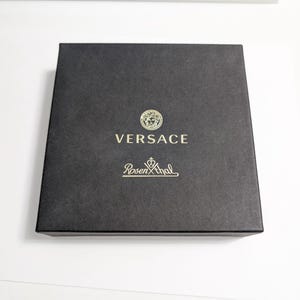 Puede incluir: Una caja cuadrada negra con el logotipo dorado de Versace, con un emblema de cabeza de Medusa, y el nombre de la marca Rosenthal. La caja tiene una superficie texturizada y est&aacute; sobre un fondo blanco.