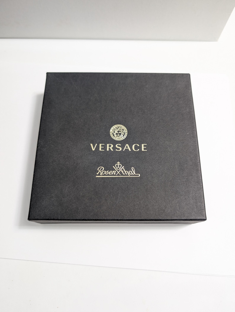 Puede incluir: Una caja cuadrada negra con el logotipo dorado de Versace, con un emblema de cabeza de Medusa, y el nombre de la marca Rosenthal. La caja tiene una superficie texturizada y est&aacute; sobre un fondo blanco.