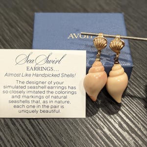 Pode incluir: Um par de brincos dourados com fechos em forma de concha e pingentes em forma de conchas espirais cor-de-rosa claro. Os brincos são apresentados com um cartão que diz: Sea Swirl EARRINGS... Almost Like Handpicked Shells! The designer of your simulated seashell earrings has so closely imitated the colorings and markings of natural seashells that, as in nature, each one in the pair is uniquely beautiful.