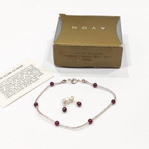 May include: A silver bracelet and earrings set with red garnet beads. The set comes in a gold box with the text "Liquid Silver Garnet Bead Gift Set" and "small". The box also includes a card with the text "The Allure of Silver" and a description of the jewelry.