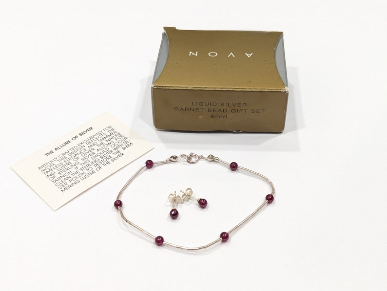 May include: A silver bracelet and earrings set with red garnet beads. The set comes in a gold box with the text "Liquid Silver Garnet Bead Gift Set" and "small". The box also includes a card with the text "The Allure of Silver" and a description of the jewelry.
