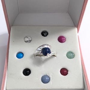 May include: A silver ring with a dark blue gemstone, presented in a pink jewelry box. The box contains the ring and several colorful gemstones, including blue, green, pink, and black.