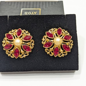 May include: A pair of gold tone earrings with a red rhinestone flower design and a pearl center. The earrings are displayed on a black velvet background with the word "AVON" printed on the box.