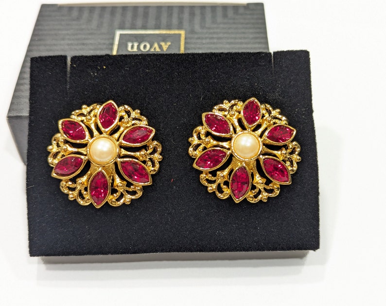 May include: A pair of gold tone earrings with a red rhinestone flower design and a pearl center. The earrings are displayed on a black velvet background with the word "AVON" printed on the box.