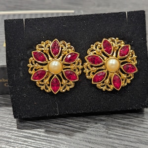 May include: A pair of gold tone earrings with a floral design. Each earring features a cluster of red rhinestones and a single pearl in the center.