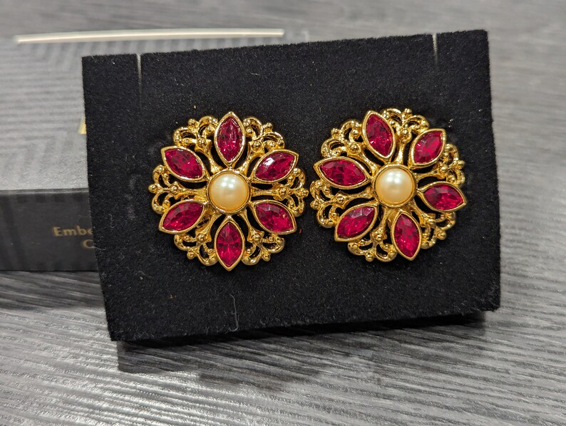 May include: A pair of gold tone earrings with a floral design. Each earring features a cluster of red rhinestones and a single pearl in the center.