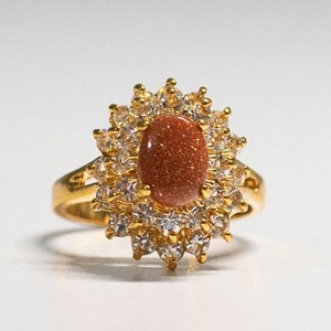 May include: A gold-toned ring featuring an oval, reddish-brown stone at the center, surrounded by a halo of clear, sparkling stones. The ring's band is a simple, polished gold. The design is reminiscent of a sunburst.