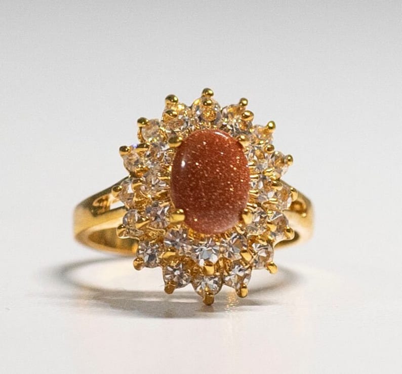 May include: A gold-toned ring featuring an oval, reddish-brown stone at the center, surrounded by a halo of clear, sparkling stones. The ring's band is a simple, polished gold. The design is reminiscent of a sunburst.