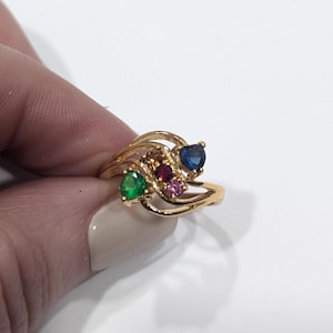 以下が含まれることがあります： 渦巻き模様のゴールドリングに、緑、赤、ピンク、青の4つのカラフルな宝石がセットされています。