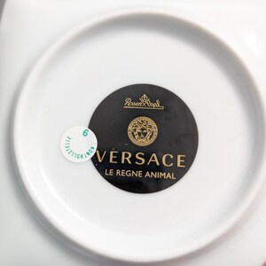 Puede incluir: Un plato de porcelana blanca con forma cuadrada y una hendidura circular. La parte inferior del plato presenta un c&iacute;rculo negro con el logotipo de Versace en dorado y el texto "LE REGNE ANIMAL". Una peque&ntilde;a pegatina es visible.