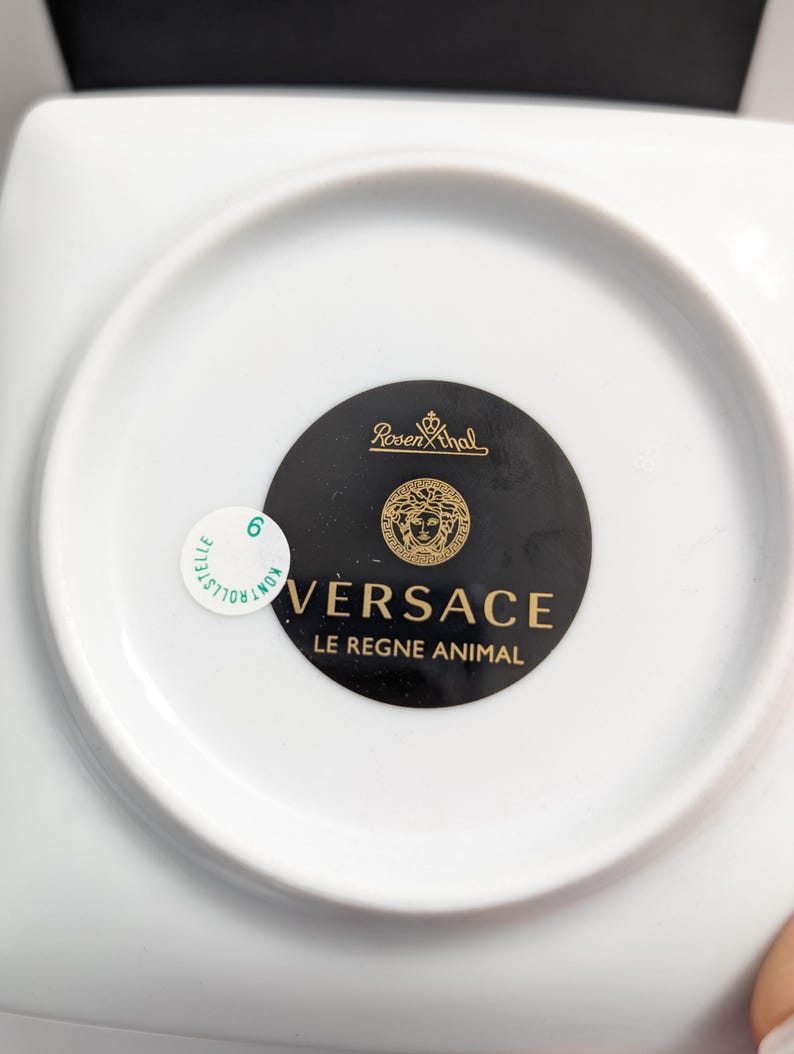 Puede incluir: Un plato de porcelana blanca con forma cuadrada y una hendidura circular. La parte inferior del plato presenta un c&iacute;rculo negro con el logotipo de Versace en dorado y el texto "LE REGNE ANIMAL". Una peque&ntilde;a pegatina es visible.