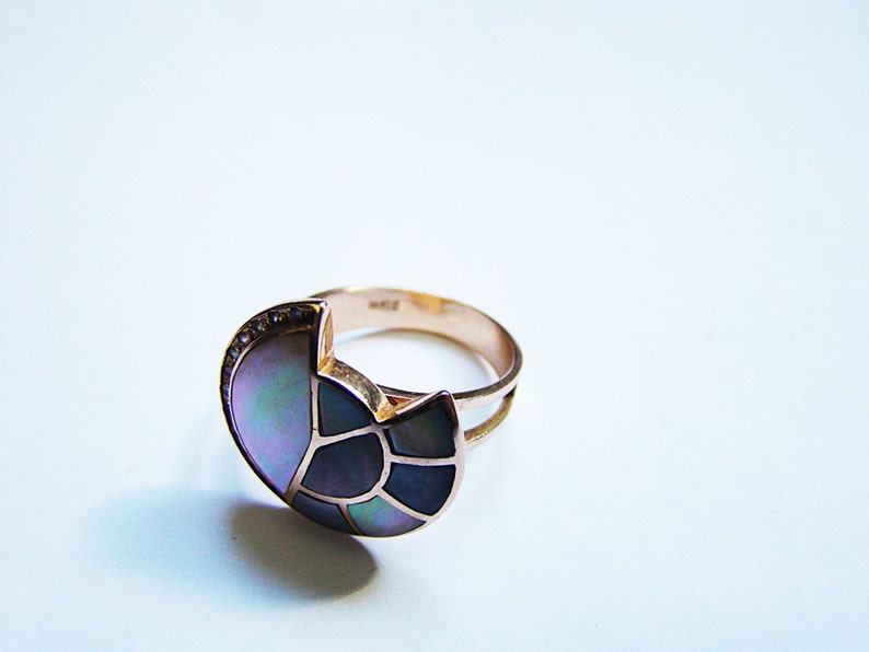 May include: A gold ring with a large, iridescent, multi-colored stone set in the center. The stone is shaped like a crescent moon and has a geometric pattern.