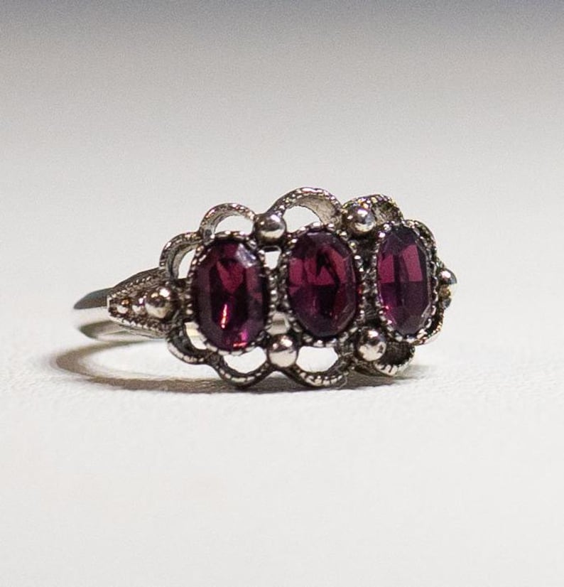 May include: A silver ring with three oval-shaped purple gemstones set in a delicate filigree design.