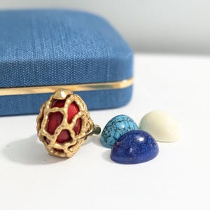 May include: A gold-coloured ring with a red stone centre and a cage-like design, beside three cabochon stones in blue and cream colours. A blue box with gold trim is in the background. Jewellery and accessories.