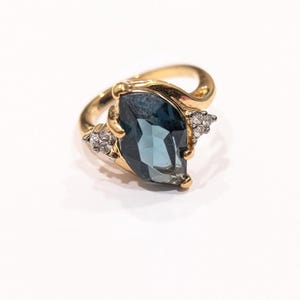 May include: A gold-toned ring featuring a large, faceted, teardrop-shaped blue gemstone. The ring has a decorative design with small, clear accent stones on either side of the main stone. The ring is set against a white background.