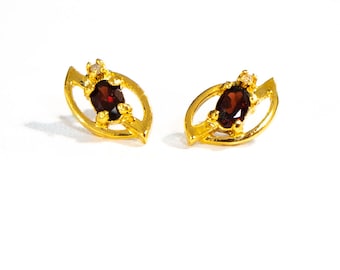 May include: A pair of gold earrings with a red gemstone and small clear stones. The earrings are shaped like a leaf.