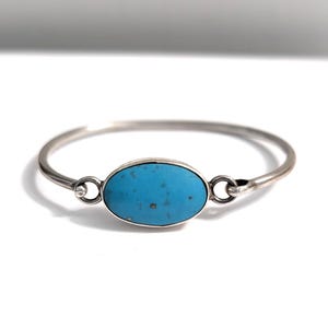 May include: A silver-toned bangle bracelet with a bright turquoise oval stone at the center. The stone is set in a silver bezel and is connected to the bracelet's band with small silver loops. The bracelet is displayed on a white background.