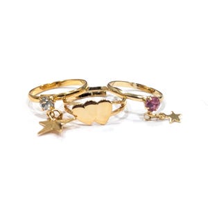 May include: Three gold-toned rings arranged together. The left ring features a clear stone and a dangling star charm. The center ring displays two connected heart shapes. The right ring includes a pink stone and a dangling star charm.