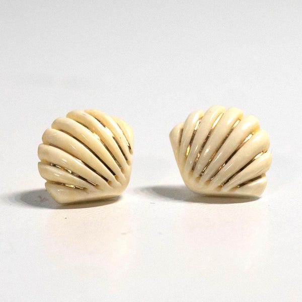 Puede incluir: Un par de pendientes de botón en forma de concha de color crema, con una textura acanalada y sutiles acentos dorados entre las crestas. Los pendientes tienen un acabado liso y pulido, con un diseño curvo en forma de abanico que imita una vieira.