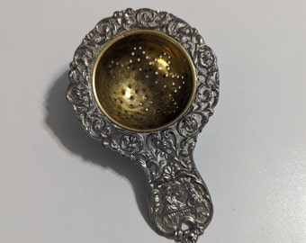 Cuchara coladora de té antigua de plata y bronce alemán con decoración: escena costera en relieve