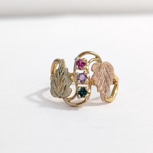 Op de afbeelding: Een goudkleurige ring met drie edelstenen in roze, paars en groen. De ring heeft bladaccenten in goud- en roségoudtinten, met een delicaat, door de natuur geïnspireerd ontwerp.