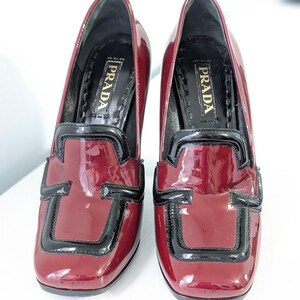 May include: Pair of burgundy patent leather heeled loafers with black trim. The shoes have a square toe and a block heel. The brand name "PRADA" is visible on the insole.