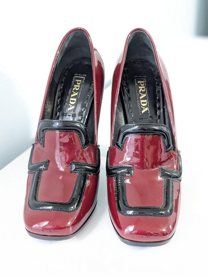 May include: Pair of burgundy patent leather heeled loafers with black trim. The shoes have a square toe and a block heel. The brand name "PRADA" is visible on the insole.