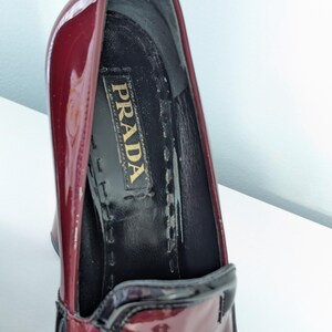 May include: Close-up of a burgundy patent leather Prada high heel shoe. The shoe features a black trim and a black interior with the Prada logo. The shoe is angled, showing the inside and the glossy exterior.