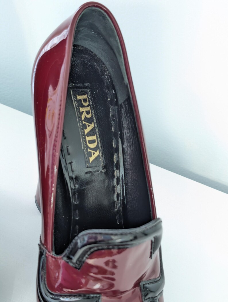 May include: Close-up of a burgundy patent leather Prada high heel shoe. The shoe features a black trim and a black interior with the Prada logo. The shoe is angled, showing the inside and the glossy exterior.