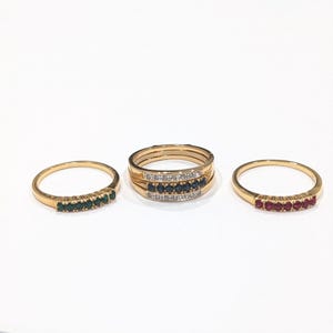 May include: Three gold rings are displayed on a white surface. One ring features a row of green gemstones, another has a row of blue gemstones and diamonds, and the third has a row of red gemstones.