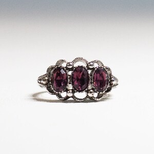 May include: A silver ring with three oval-shaped purple gemstones set in a delicate filigree design.