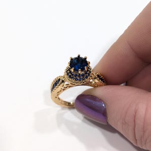 Peut inclure: Une bague en or avec une grande pierre pr&eacute;cieuse bleue entour&eacute;e de petites pierres bleues. La bague a un design d&eacute;licat et complexe.