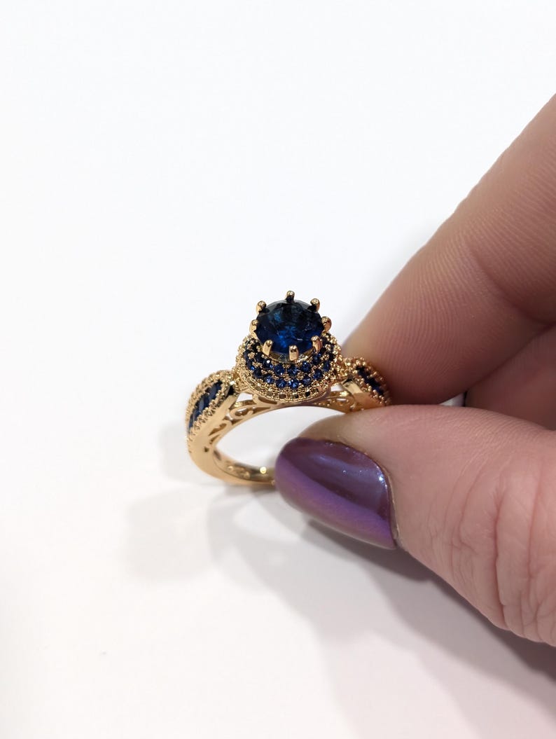 Peut inclure: Une bague en or avec une grande pierre pr&eacute;cieuse bleue entour&eacute;e de petites pierres bleues. La bague a un design d&eacute;licat et complexe.