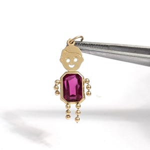 Peut inclure: Un pendentif en or en forme de personnage avec un visage souriant, doté d'une pierre précieuse rectangulaire à facettes rose foncé en guise de torse, avec de petites sphères en or formant les bras et les jambes, tenu par une pince.