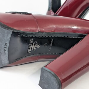 May include: Close-up of a pair of burgundy patent leather high-heeled shoes. The soles are black with the "PRADA" logo and "MADE IN ITALY" text. The heel is a block style.