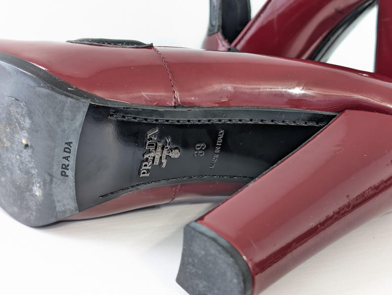 May include: Close-up of a pair of burgundy patent leather high-heeled shoes. The soles are black with the "PRADA" logo and "MADE IN ITALY" text. The heel is a block style.