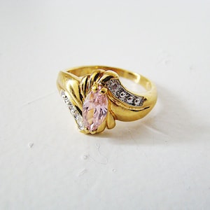 May include: A gold ring with a pink marquise-shaped gemstone surrounded by smaller clear gemstones. The ring has a swirl design on the band.