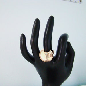 May include: A ring with a large, white, geometric design set in gold. The ring is on a black hand model.