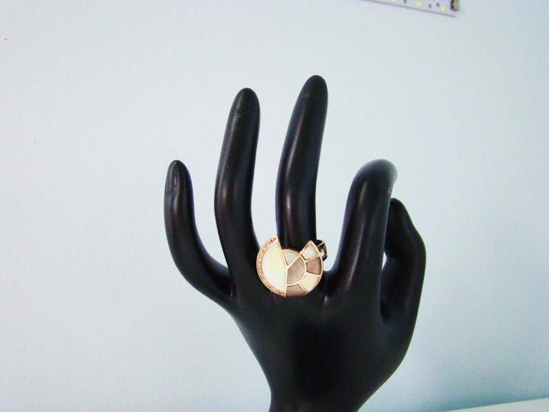 May include: A ring with a large, white, geometric design set in gold. The ring is on a black hand model.