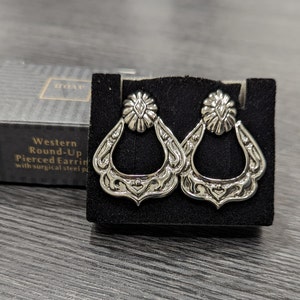 May include: A pair of silver Western Round-Up Pierced Earrings with a detailed, ornate design. The earrings are in a black velvet box with the words "Western Round-Up Pierced Earrings with surgical steel posts" printed on the box.