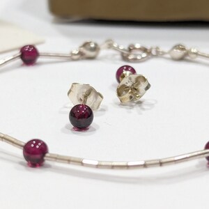 May include: A silver chain bracelet with small, round, dark red gemstones. The bracelet is paired with matching stud earrings with dark red gemstones.