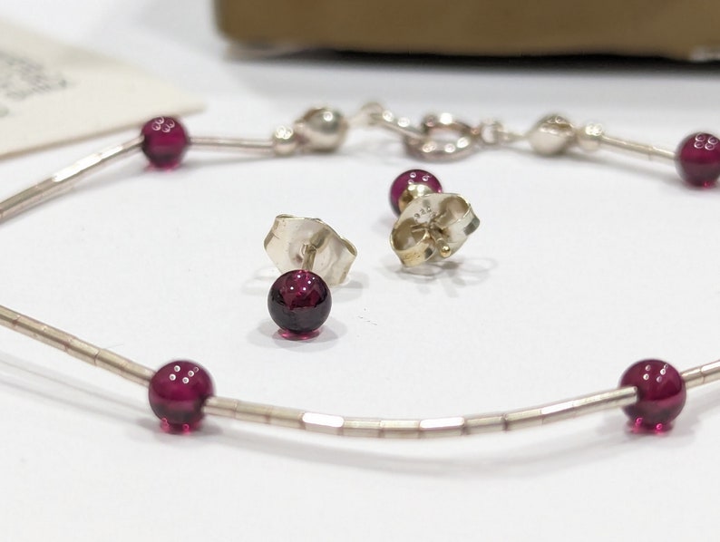 May include: A silver chain bracelet with small, round, dark red gemstones. The bracelet is paired with matching stud earrings with dark red gemstones.