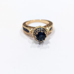 Peut inclure: Une bague en or avec une grande pierre pr&eacute;cieuse bleue entour&eacute;e de petites pierres bleues. La bague a un design d&eacute;licat et complexe.