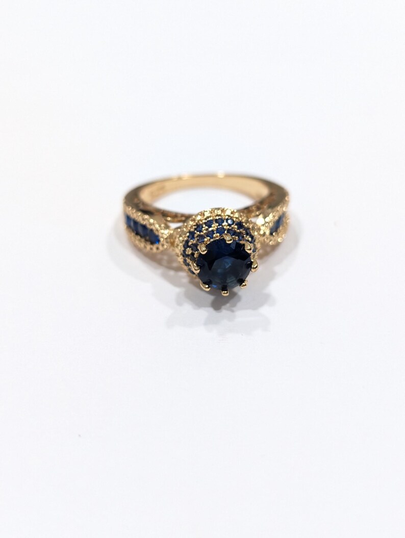 Peut inclure: Une bague en or avec une grande pierre pr&eacute;cieuse bleue entour&eacute;e de petites pierres bleues. La bague a un design d&eacute;licat et complexe.
