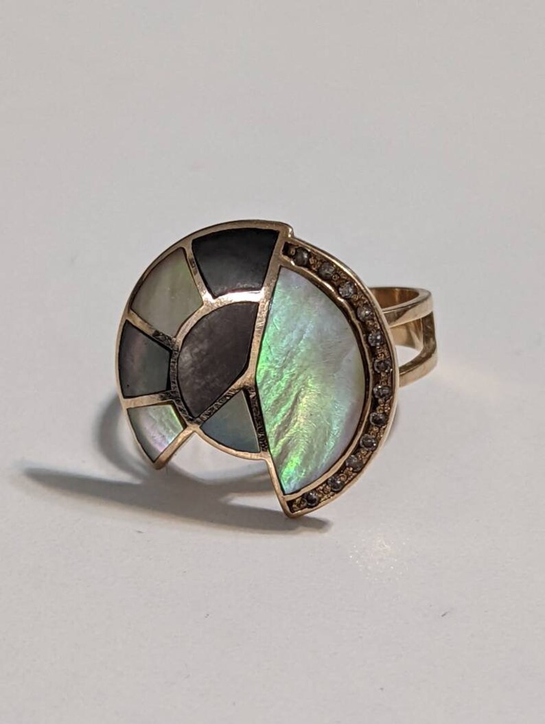 May include: A gold ring with a large, multi-colored, iridescent, abalone shell design. The shell is set in a gold band and has a row of small diamonds around the edge.