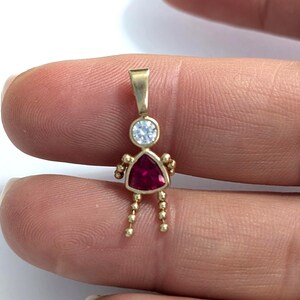 May include: A gold pendant in the shape of a stick figure. The head is a round, clear stone, and the body is a triangular, red stone. The arms and legs are made of small gold beads. The pendant is held between two fingers.
