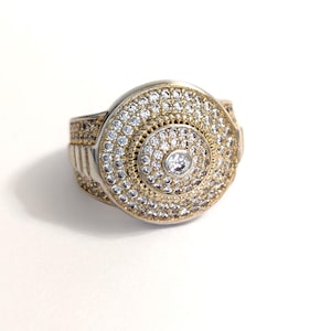 Puede incluir: Un anillo de plata y oro con una gran cara circular. La cara está cubierta de pequeñas piedras preciosas transparentes, con una piedra preciosa más grande en el centro. La banda tiene un diseño texturizado.