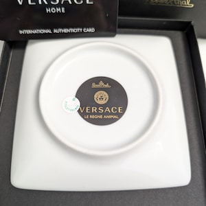 Puede incluir: Un plato cuadrado de porcelana blanca con una hendidura circular en el centro. El plato tiene el texto "VERSACE LE REGNE ANIMAL" y un emblema dorado. Se encuentra en una caja negra con una tarjeta de autenticidad Versace Home.